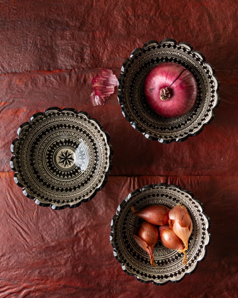 Three hand-painted Moroccan ceramic bowls with scalloped edges and black and white geometric designs, holding a red onion and shallots.