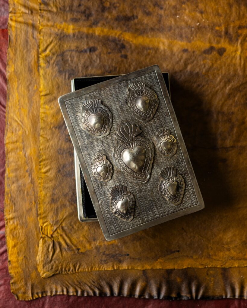 A hand-tooled silver Milagro keepsake box lid, embossed with seven sacred hearts, resting on an open velvet-lined box over yellow and red-brown leather.