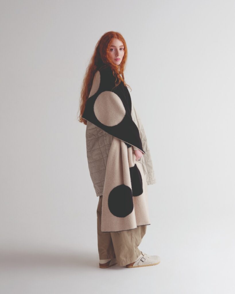 A handmade chunky knit throw blanket in a black and beige polka dot pattern being modeled by a woman with red hair, over a quilted jacket.