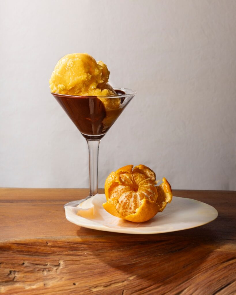 A martini glass filled with orange citrus sorbet melting into chocolate sauce, next to a partially peeled clementine on a mother-of-pearl plate.