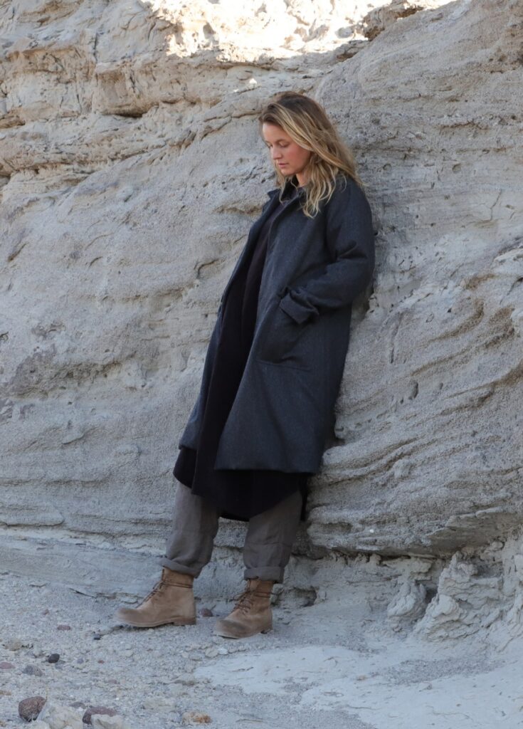 A woman models a long, dark gray Italian wool coat with corozo nut buttons, posing against a pale, rocky cliffside.
