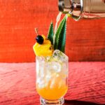 A Jungle Bird tiki cocktail being poured from a shaker, garnished with a pineapple wedge, cherry, and fronds, on a vibrant red wooden surface.