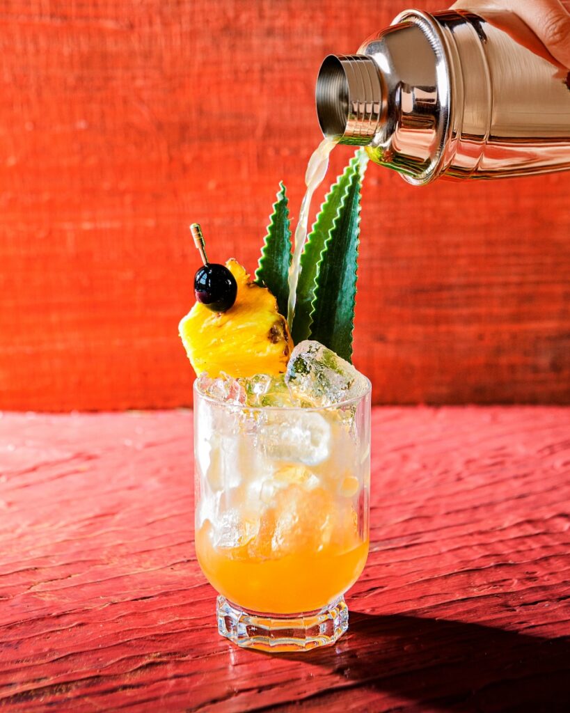 A Jungle Bird tiki cocktail being poured from a shaker, garnished with a pineapple wedge, cherry, and fronds, on a vibrant red wooden surface.