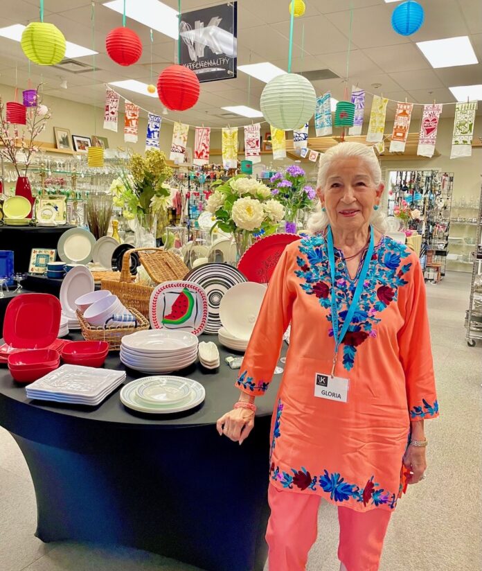 A woman in pink and blue stands beside a table.