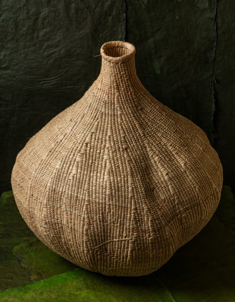 A large, handwoven 'garlic' shaped storage basket made from natural ilala palm grasses by artisans in Zimbabwe, sitting on a green surface.