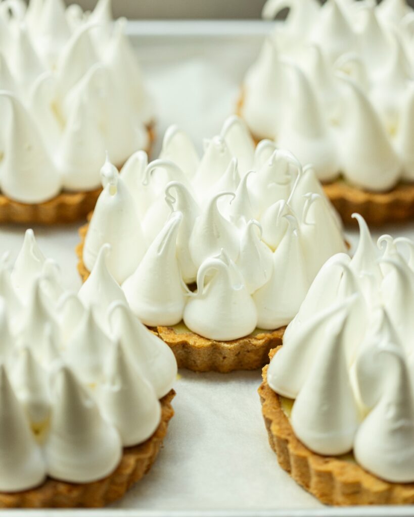 Mini lemon meringue tarts with tall, stiff, white piped meringue peaks on a white surface.