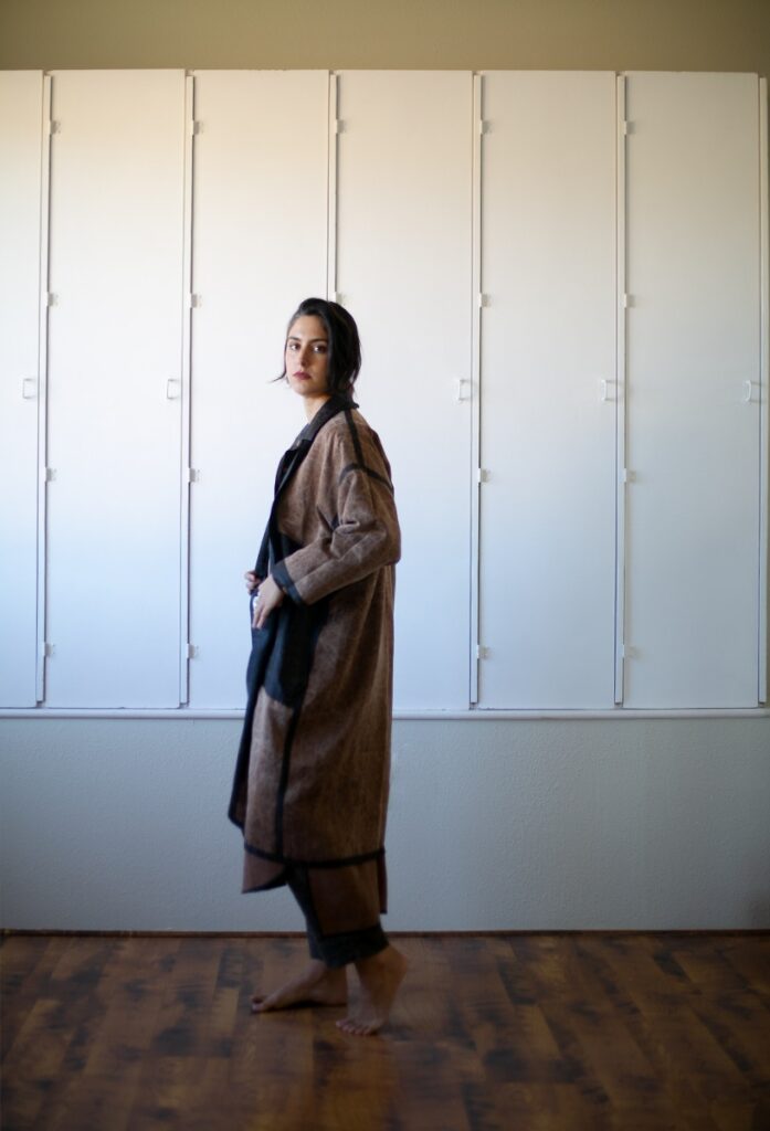 A woman modeling a long, draped, two-tone brown and black coat made from luxurious Guangdong mud silk, standing in front of a white paneled wall.