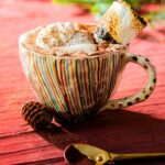 A multicolored striped ceramic mug of Mexican Hot Chocolate topped with whipped cream and a toasted marshmallow, next to a gold spoon and pine cone, on a red wooden surface.