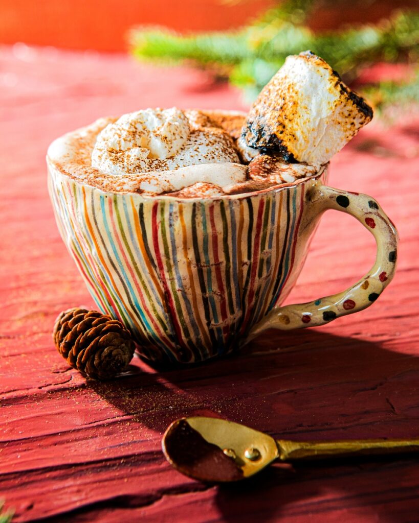 A multicolored striped ceramic mug of Mexican Hot Chocolate topped with whipped cream and a toasted marshmallow, next to a gold spoon and pine cone, on a red wooden surface.