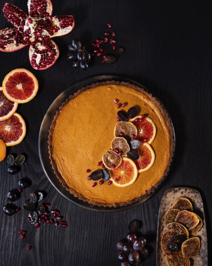 A miso sweet potato pie sits on a dark wood table with sliced citrus, grapes, and pomegranate seeds on top.