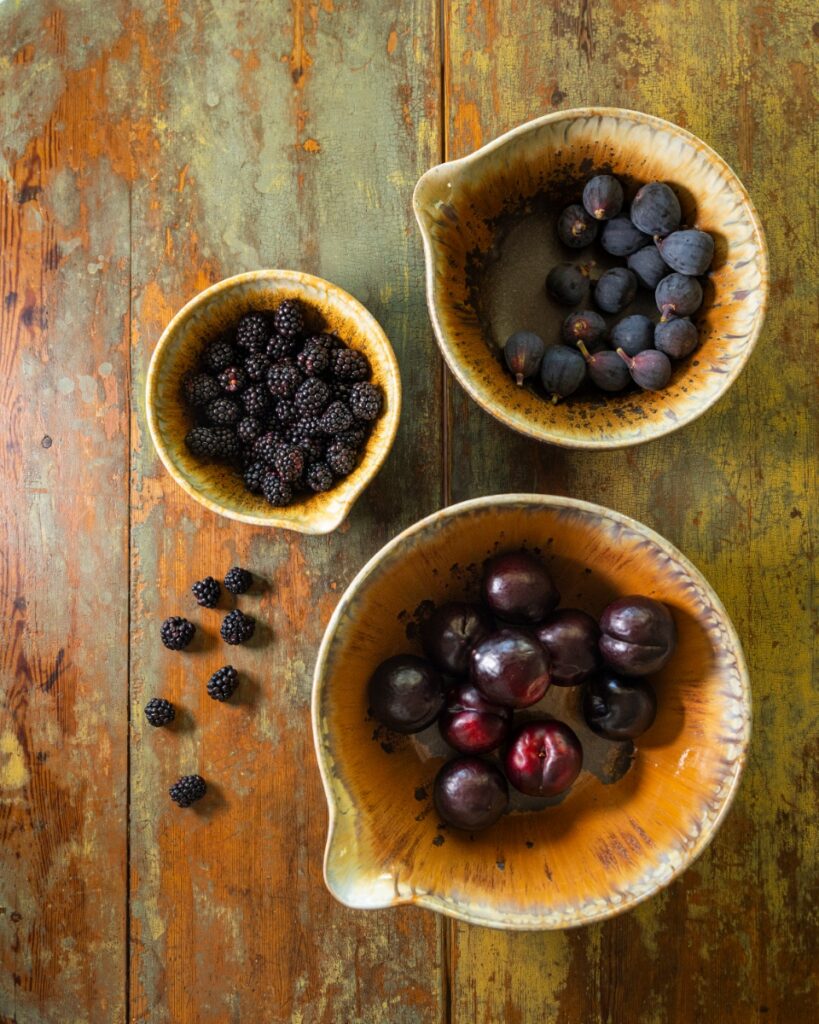 Three nested stoneware bowls with a warm orange-brown glaze, filled with fresh dark plums, figs, and blackberries, on a distressed wood table.