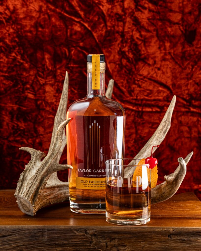 A bottle of Taylor Garrett Old Fashioned cocktail next to a garnished glass, flanked by two antlers on a dark wood table.