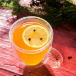 A clear glass mug containing a warm orange Pear Hot Toddy, garnished with a clove-studded lemon slice, set on a red wooden background with pine needles.