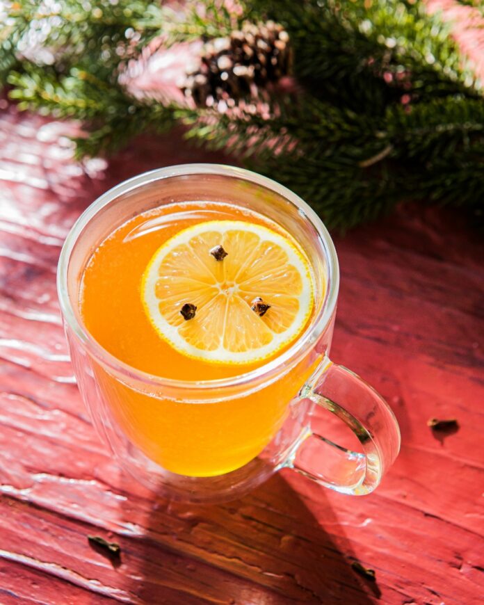 A clear glass mug containing a warm orange Pear Hot Toddy, garnished with a clove-studded lemon slice, set on a red wooden background with pine needles.