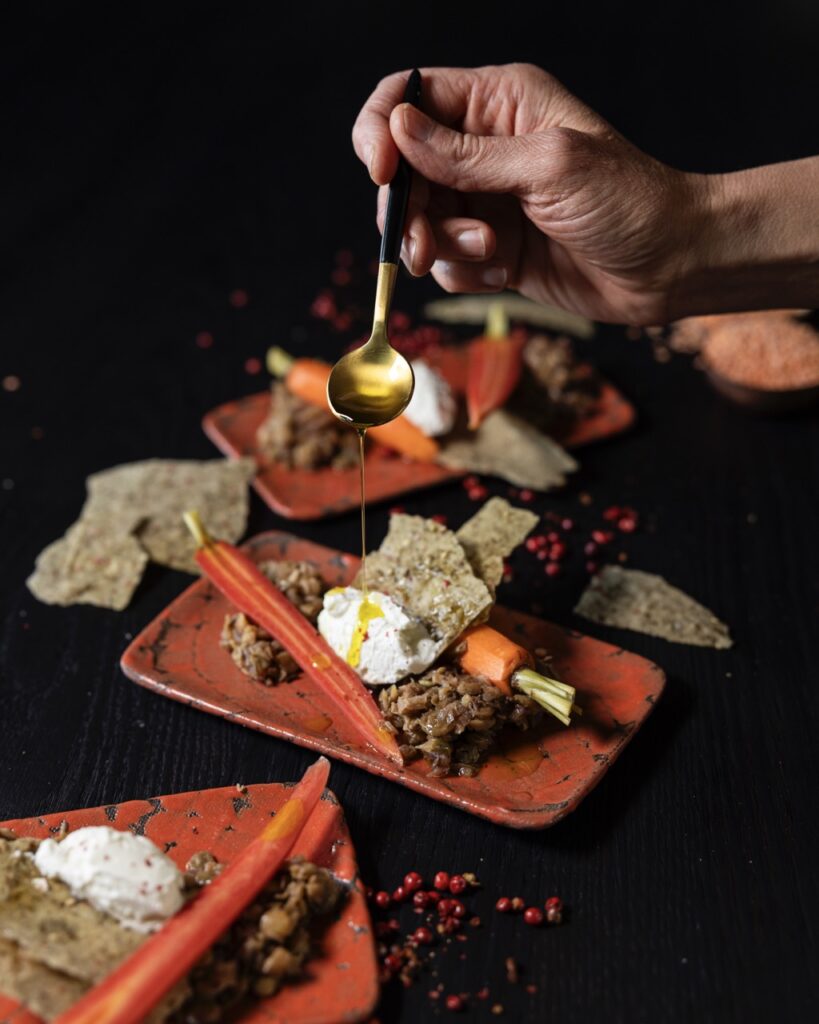 A person drizzles honey over a small orange plate of pickled baby carrots with smoked labneh and lentils.