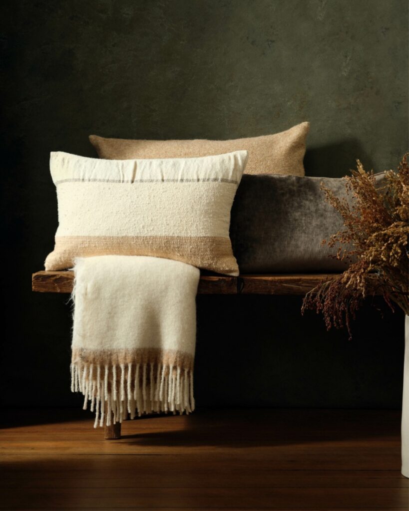 A natural-toned collection of pillows and a fringed blanket made from organic materials and fibers like wool and linen, resting on a wooden bench.