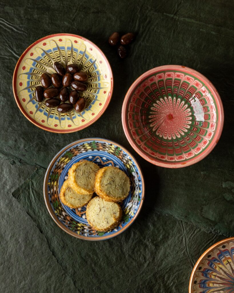Three pieces of brightly hand-painted Romanian Horezu pottery bowls and plates, filled with olives and small butter cookies, on a dark surface.