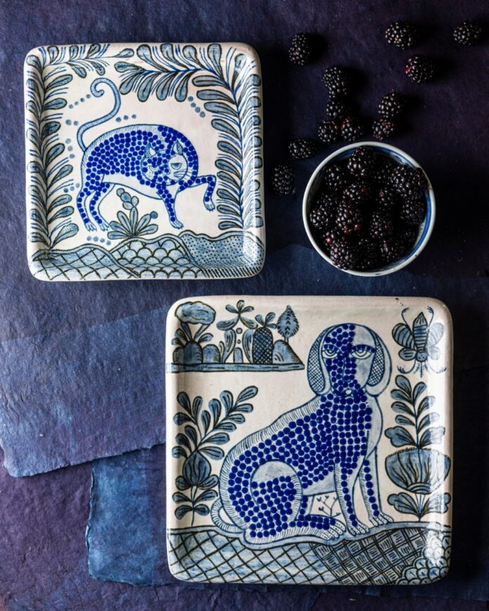 Two square ceramic plates with hand-painted blue and white whimsical folk art designs featuring a dancing cat and a sitting dog, beside a bowl of blackberries.
