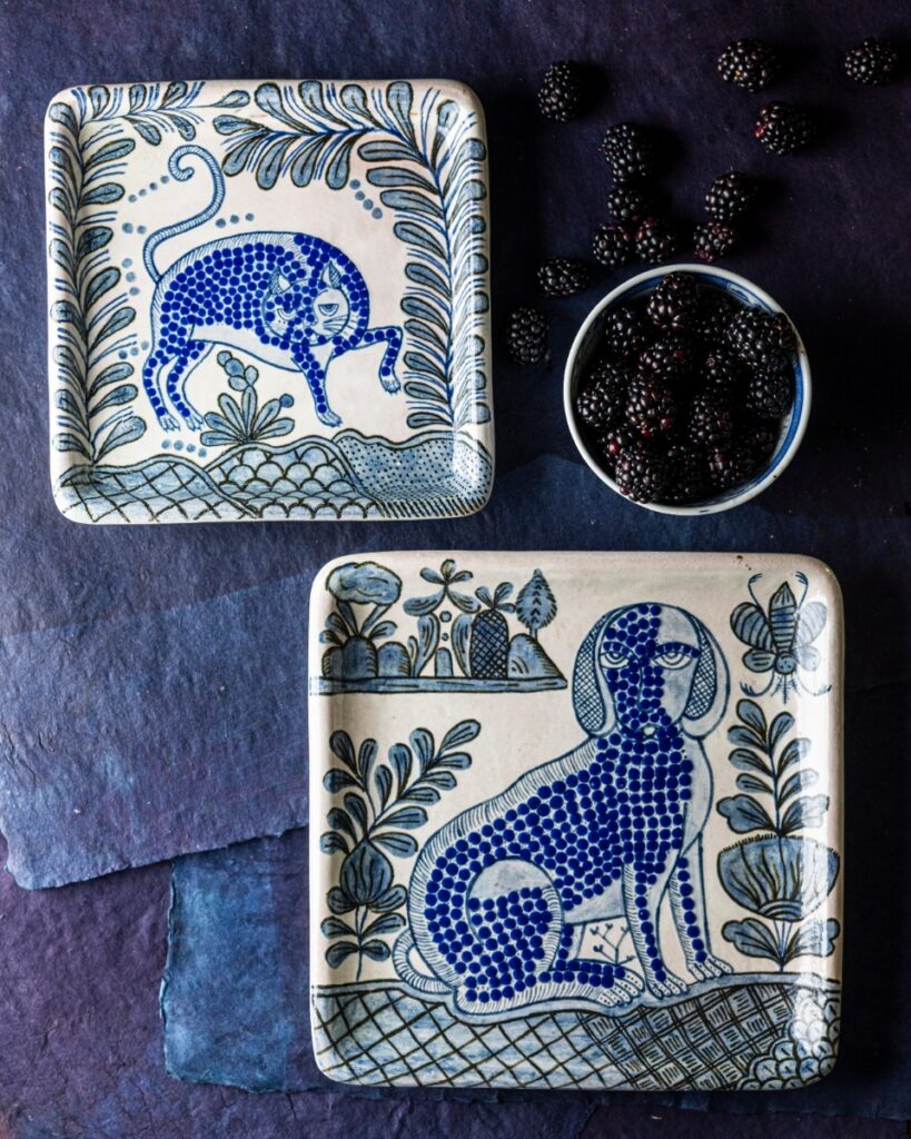 Two square ceramic plates with hand-painted blue and white whimsical folk art designs featuring a dancing cat and a sitting dog, beside a bowl of blackberries.