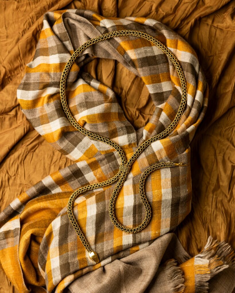 A gold 18K-dipped beaded snake necklace lying on a plaid cashmere hand-loomed shawl in mustard yellow, white, and brown.