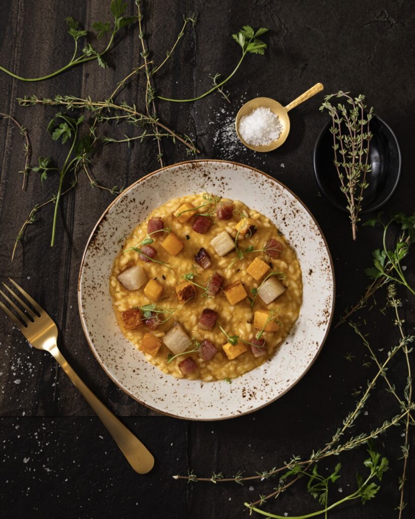 A white plate with an Apple Cider Risotto in an orange color from The Compound Restaurant sits on a table with springs of greenery.