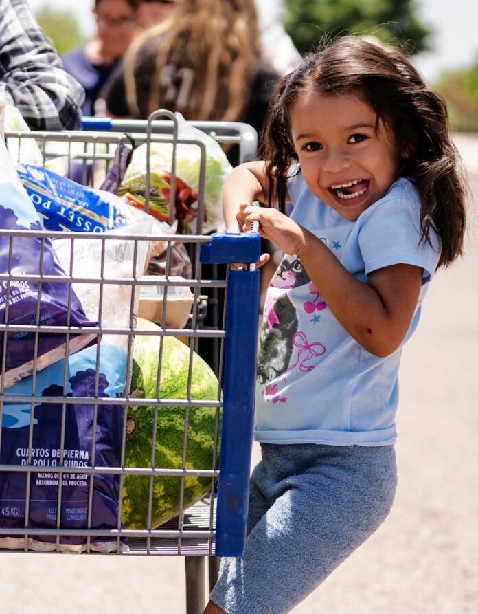 A little girl holds on and rides the side of a shopping cart.