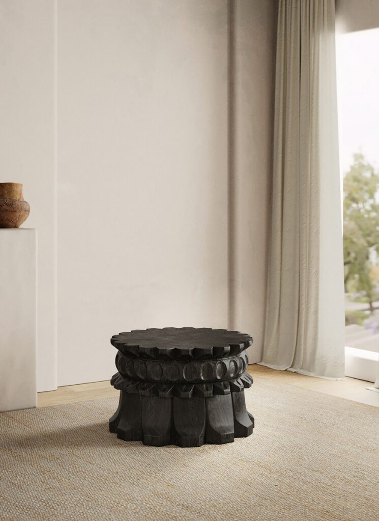 A dark, carved wooden Molinillo cocktail table with a turned base and circular motif, next to sheer curtains and a light wall in a modern living room.