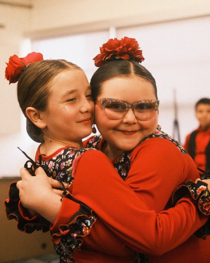 Two girls in red costumes and hair pieces hug each other.