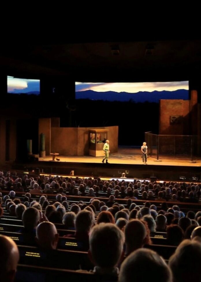 People sit in an outside theater with opera actors on a stage.
