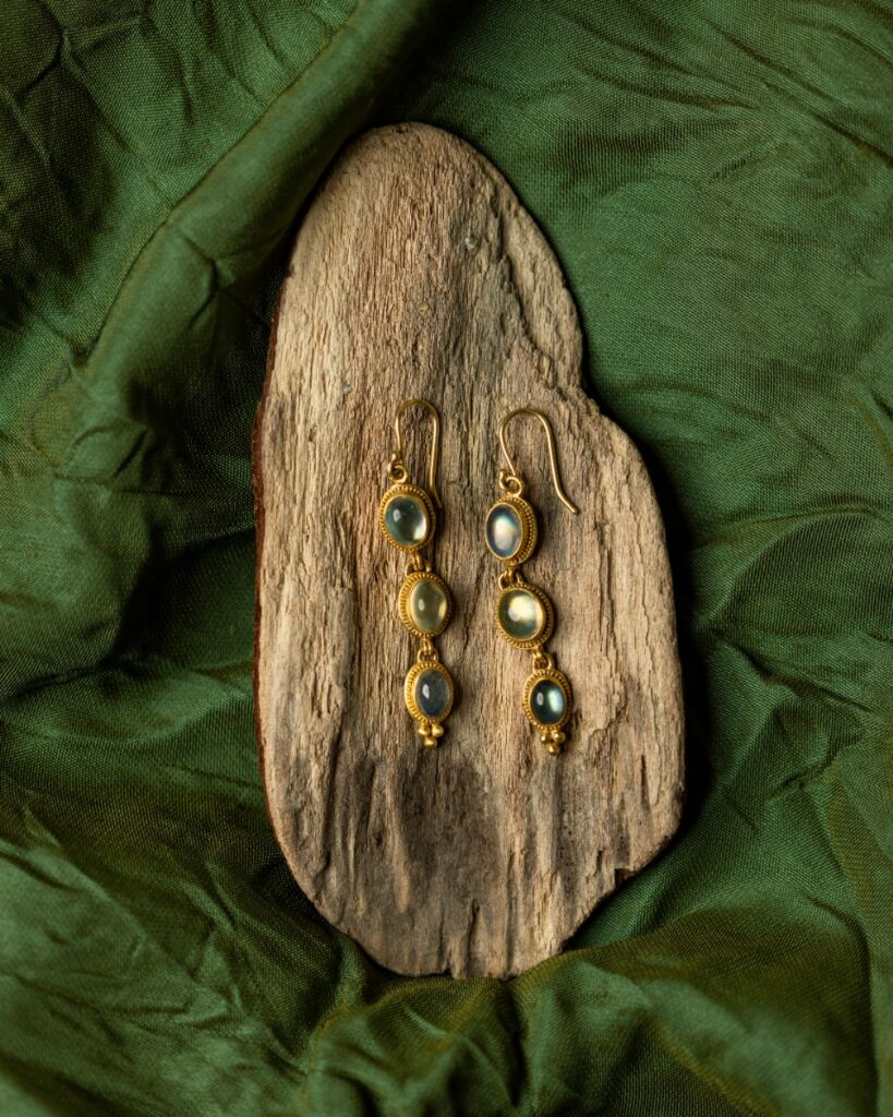 Gold earrings by Luna Felix with three hinged moonstone cabochons and granulation details, resting on a piece of driftwood and a dark green cloth.