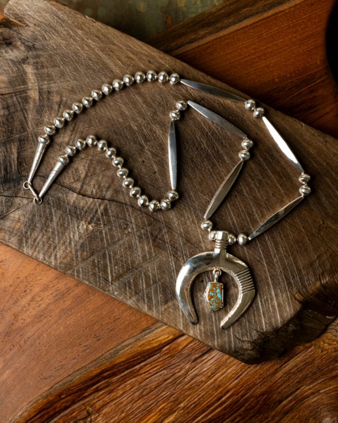 Tufa cast sterling silver necklace by Cippy Crazy Horse with Navajo pearls, a heavy-gauge Naja pendant, and a small turquoise stone hanging on rustic wood.