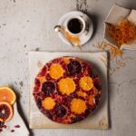 A round, vibrant upside-down citrus cake topped with sliced oranges, blood oranges, and pomegranate seeds, next to a cup of coffee.