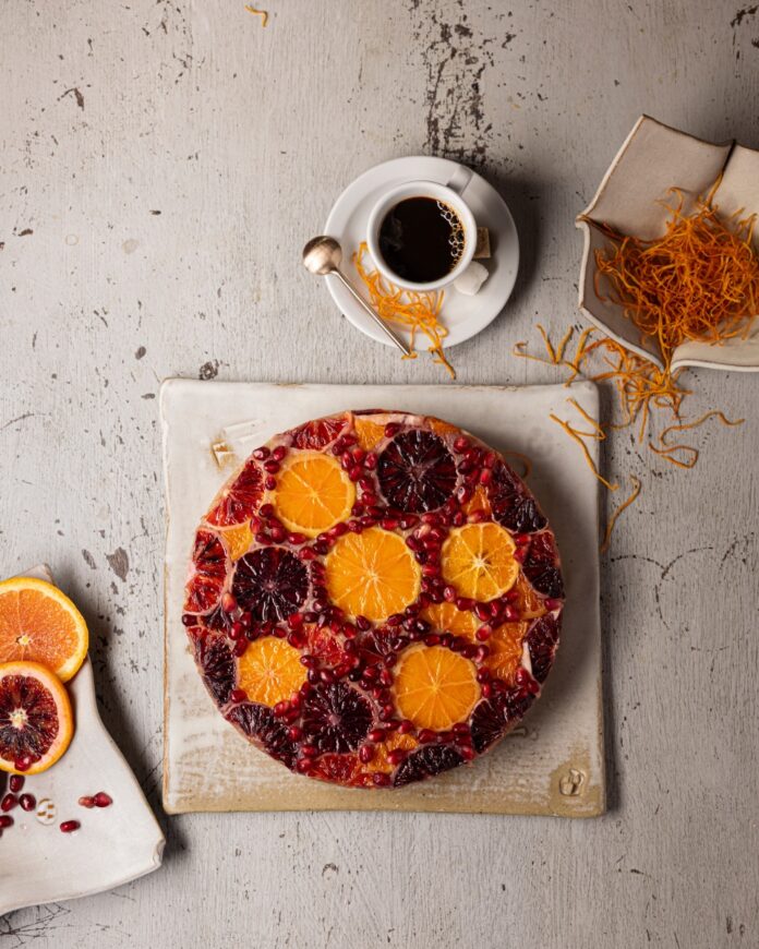 A round, vibrant upside-down citrus cake topped with sliced oranges, blood oranges, and pomegranate seeds, next to a cup of coffee.