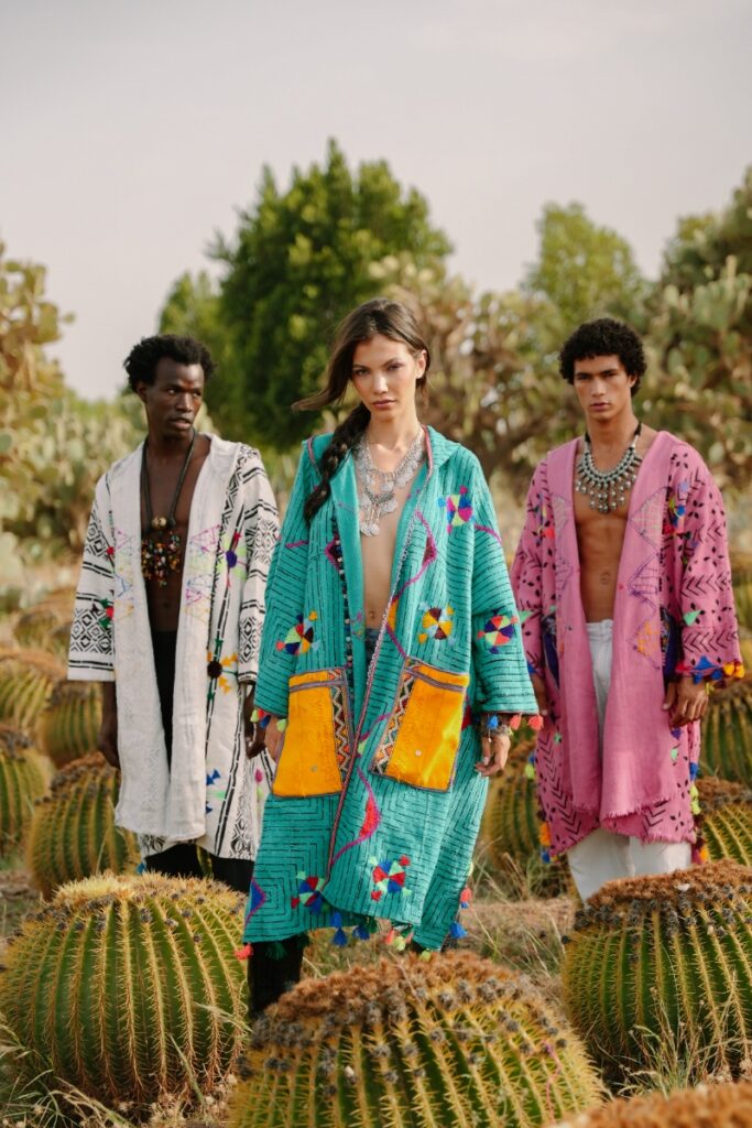 Three models wearing colorful, handwoven, and embroidered Moroccan Saharan coats over simple clothing, standing among spherical cactus plants.