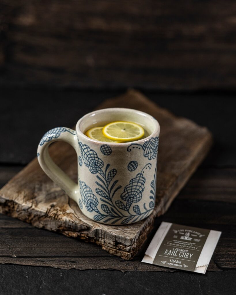 A rustic ceramic mug from Arte Ventosa with a blue floral pattern, filled with Lavender Earl Grey tea and lemon slices, on a wooden board.
