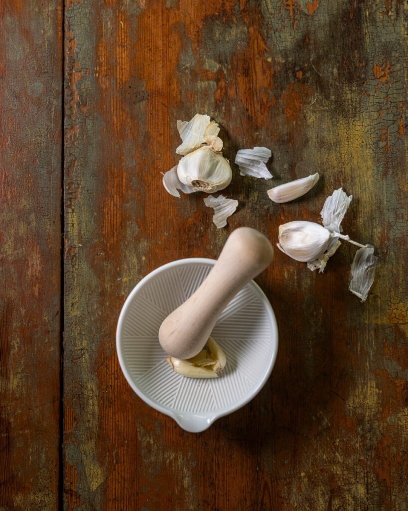 A small white porcelain mortar with a wooden maple pestle and garlic cloves on a rustic distressed wooden surface.