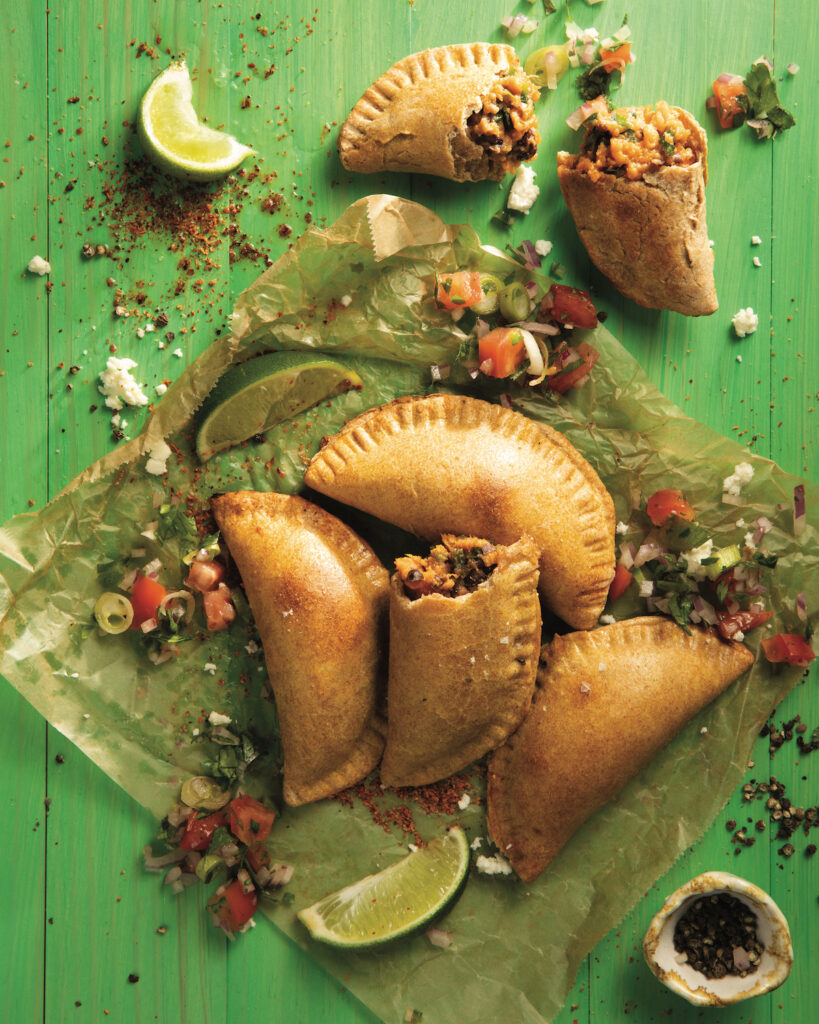Five homemade Empanadas sit on green wax paper. the Empanada in the middle has half of it eaten. homemade Empanadas recipe