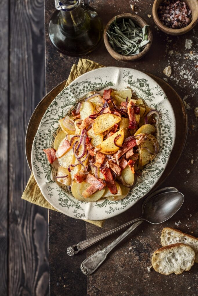 A plate of Ampezzo-style potatoes (sliced potatoes) with crispy speck and red onions, topped with black pepper and served on a patterned rustic plate with silver spoons and bread.