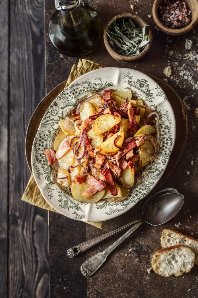 A plate of Ampezzo-style potatoes (sliced potatoes) with crispy speck and red onions, topped with black pepper and served on a patterned rustic plate with silver spoons and bread.