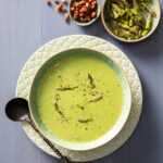 A green soup sits in a bowl with asparagus spear heads throughout the soup and in a bowl above the soup.