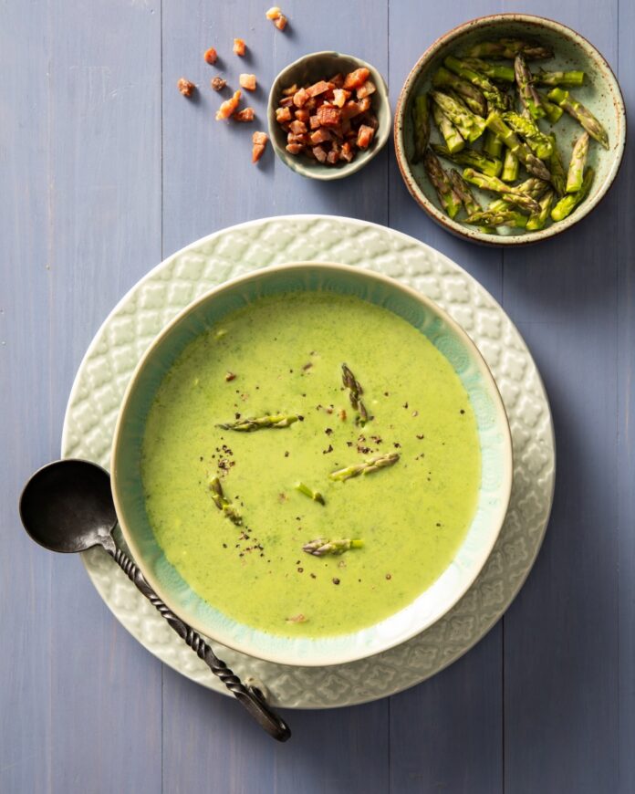 A green soup sits in a bowl with asparagus spear heads throughout the soup and in a bowl above the soup.