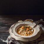 A bowl of rustic barley soup with carrots, spinach, and meat, served in a decorative white-rimmed bowl on dark wood, with a spoon resting in the bowl.
