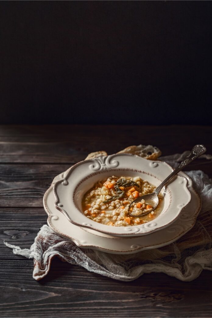 A bowl of rustic barley soup with carrots, spinach, and meat, served in a decorative white-rimmed bowl on dark wood, with a spoon resting in the bowl.