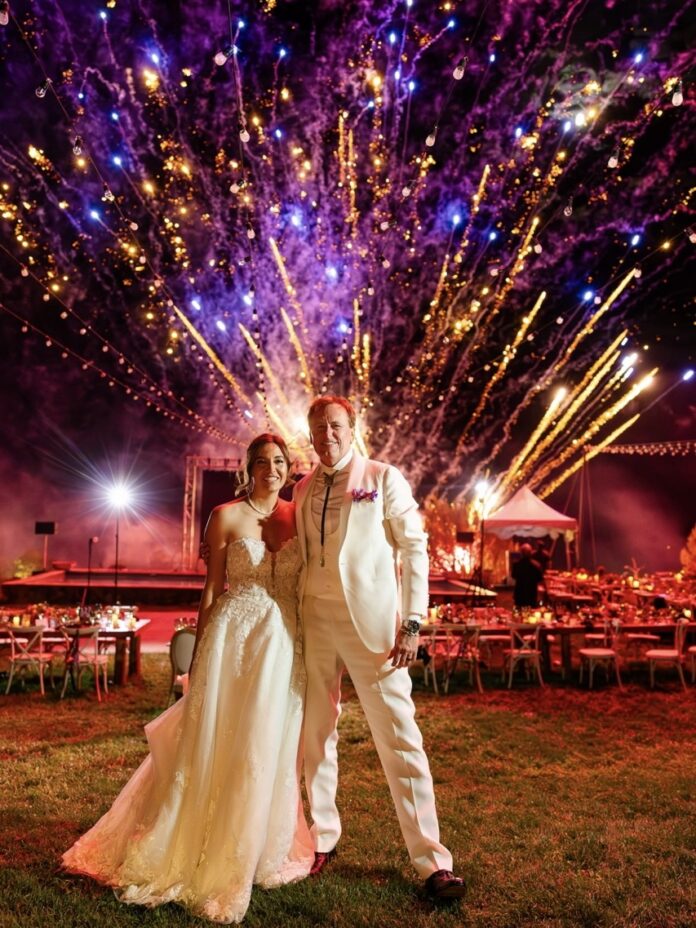 A man and woman dressed for their wedding standing in from of Blame Her Ranch with fireworks in the background.
