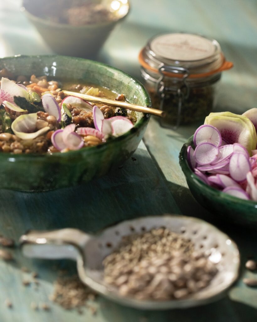 A green bowl of Bolita Bean and Fennel Sausage Soup sits in the back with a jar of herbs and a plate of beans in front of the soup.
