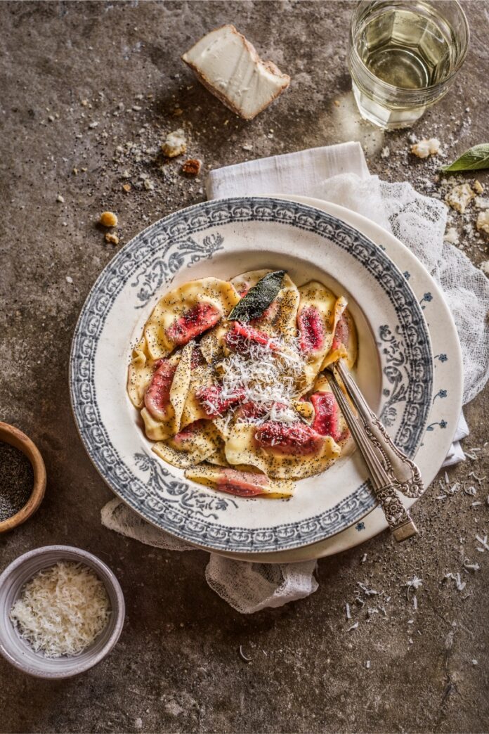 A plate of Casunziei Rossi (red beet ravioli) topped with grated parmesan and poppy seeds, garnished with a sage leaf and served with a fork on a patterned white and blue plate.