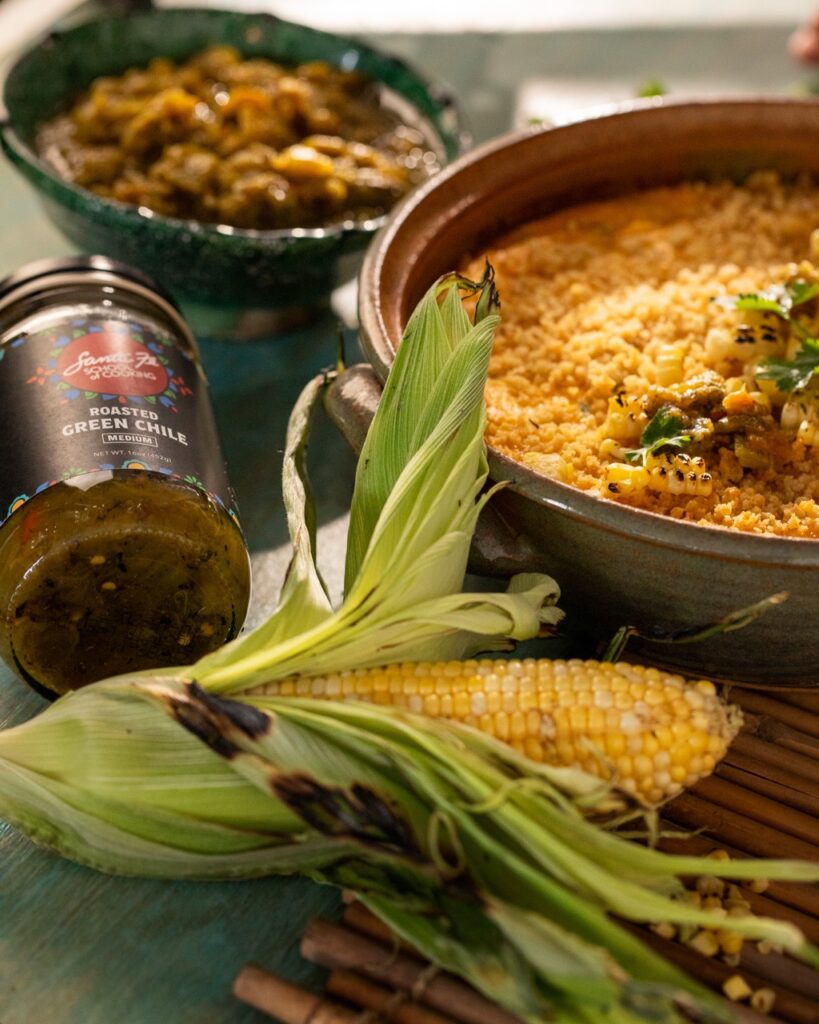 A cob of corn sits in front of a Corn Flan, jar of chiles, and bread crumbs in the back.