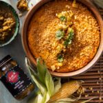 A corn flan sits in a round pan with a jar of chiles and an ear of corn below it.