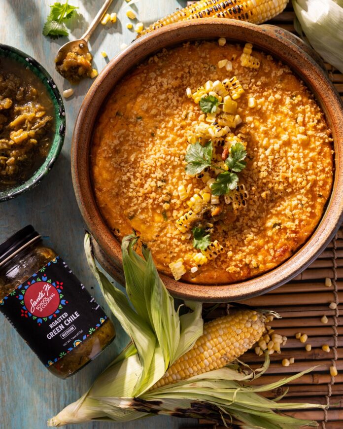 A corn flan sits in a round pan with a jar of chiles and an ear of corn below it.