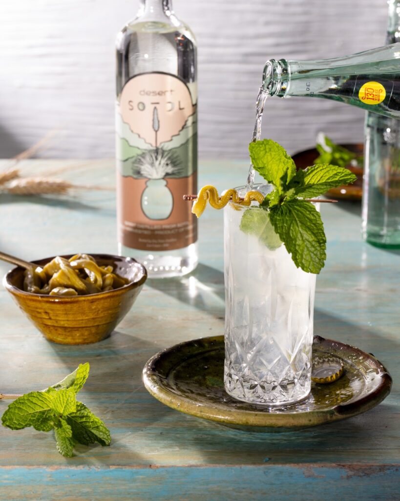 A person pours sparkling water into a glass with a mint garnish and lemon twist as a bottle of sotol sits in the background.